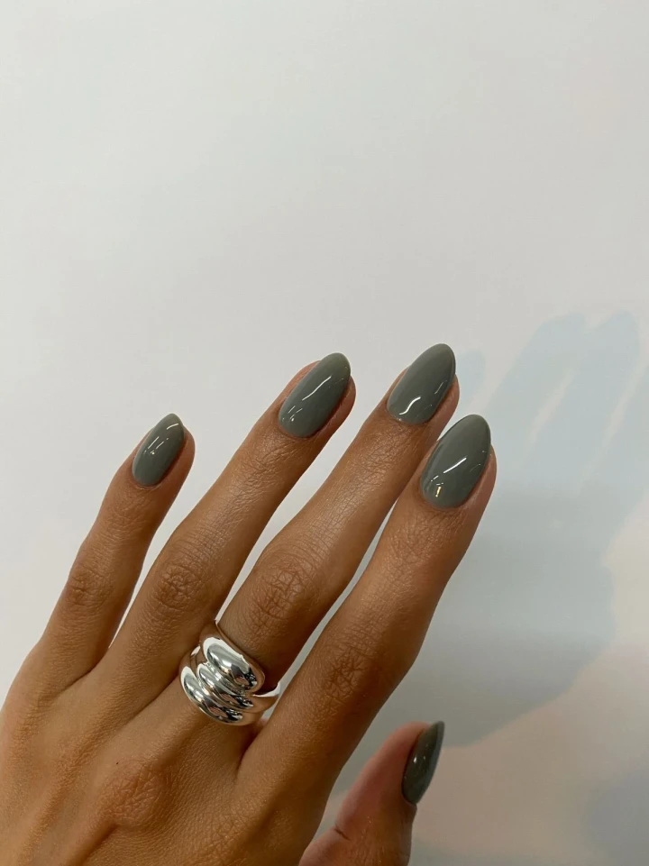 A hand with perfectly manicured nails showcasing a greyish, earthly green nail polish color, adorned with exquisite shimmering silver jewelry.
