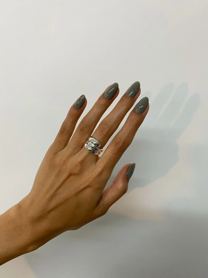 A hand with perfectly manicured nails showcasing a greyish, earthly green nail polish color, adorned with exquisite shimmering silver jewelry.